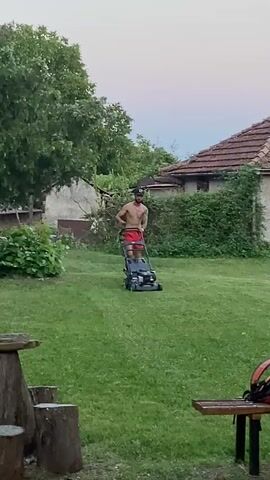 A shirtless man with tattoos is pushing a lawnmower across a grassy yard. He is wearing red shorts and a black baseball cap. The yard is surrounded by trees and bushes, and a house is visible in the background. The sky is overcast.