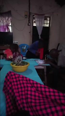 The video shows an indoor scene, likely a home or communal room, in Kiminini, Kenya. The room is dimly lit, suggesting it is evening or night. The walls are light-colored and appear to be unpainted or minimally finished.

Several children are present. In the foreground, a table covered with a light blue cloth holds a yellow bowl containing leaves or herbs, and a pink and black plaid blanket. Initially, one child, wearing a blue and red top, stands and leans over the table, seemingly looking at papers or books. Another child in a grey shirt is seated at the table on the right, actively writing in a notebook with a pen. A third child is partially visible on the far left, also seated.

As the video progresses, the child in the blue and red top moves from standing to sitting on a couch next to the child in the grey shirt. They appear to be looking at a notebook together. All the children seem engaged in their activities, which resemble studying or doing homework. Towards the end of the video, two of the children look towards the camera and smile. A blue plastic chair is visible on the left side of the table, and a television set is on a stand in the background. A door with a simple white curtain hangs in the background. Wires and a small yellowish device are visible on the wall. The overall atmosphere suggests a casual and focused domestic setting.