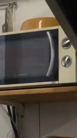 A person opens a microwave with two dials on the right side and a silver handle, removes a white-and-silver wicker basket from the interior, and pulls it out. The microwave is old and sits on a wooden shelf over white cabinets. There is an orange bowl on top of the microwave.