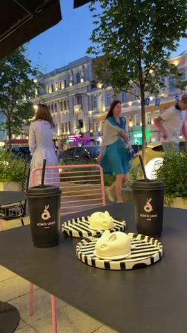 This is a video clip taken at an outdoor cafe table in Voronezh, Russia. The table has a black surface with pink legs. On the table, there are two black coffee cups with a logo that says "PONCH LIGHT CAFE" and a stylized pink bunny. There are also two small, white, bunny-shaped cakes on black and white striped plates.

In the background, there is a street scene with cars and buildings. The sky is a twilight blue, suggesting it is either early morning or late evening. One person, a woman in a blue dress, is walking away from the camera in the middle ground. Another person, a woman in a light-colored dress, is walking towards the camera in the background. The overall mood appears relaxed and pleasant, possibly indicating a casual outing or a break at a cafe.