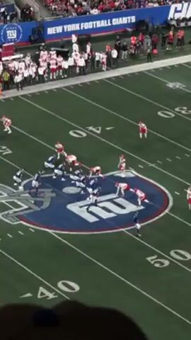 The video captures an American football game, likely between the New York Giants and the Kansas City Chiefs, from an elevated vantage point within the stadium. The teams are lined up on the field, the Giants in blue and the Chiefs in red. The play is about to begin. A large crowd of spectators fills the stands. The stadium has a green artificial turf field with yard lines and the Giants' logo prominently displayed at the center. Banners with the team name "NEW YORK FOOTBALL GIANTS" are visible above the stands.