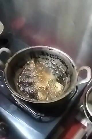 A close-up, high-angle shot shows food, possibly fish, being deep-fried in a pot of bubbling oil on a stovetop. The oil is vigorously churning around the food, creating a frothy surface. The pot is metal and has two handles. The stovetop appears to be dark. The background is blurred but suggests a kitchen environment. The lighting is artificial and somewhat dim.