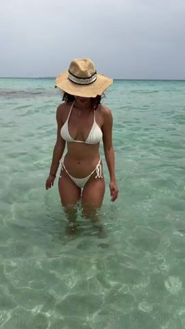 A woman in a white bikini and a straw hat stands in shallow, clear turquoise water. The water is calm with sunlight creating patterns on the sandy bottom. The woman is facing the camera and slowly walks forward, her body slightly angled to the left. She raises her right hand and waves briefly before continuing to walk. The background shows more water and a hazy horizon.