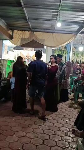 A video shows a group of people at what appears to be a gathering or celebration. The floor is covered with reddish hexagonal tiles, and the ceiling has a metal roof structure. Two women in long dresses are dancing in the foreground, facing each other. The woman on the left wears a dark red sleeveless dress, and the woman on the right wears a dark dress with off-the-shoulder sleeves. Both have long dark hair. A man in a blue t-shirt and shorts walks between them. In the background, there's a crowd of adults and children watching, some sitting on green plastic chairs. There are white and light blue curtains draped in the background, and decorative elements suggest a festive occasion, possibly a wedding. Bright lights illuminate the area.