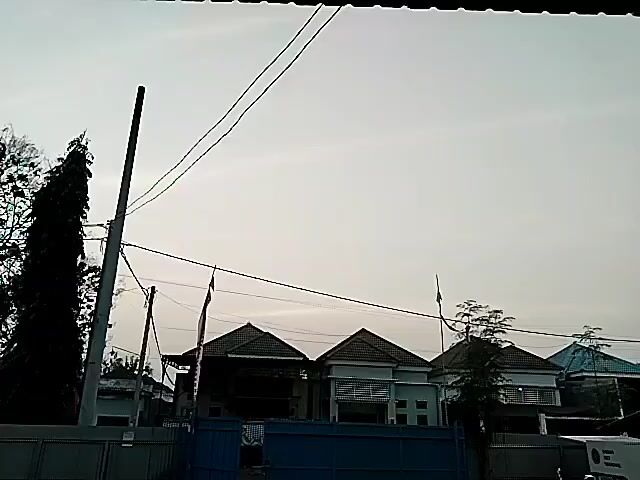 The video shows a row of houses with pitched roofs. A utility pole with wires runs through the scene. A blue gate is in the foreground. A white van with text and a logo is parked to the right. There are some trees and foliage visible. The sky is bright and overcast.