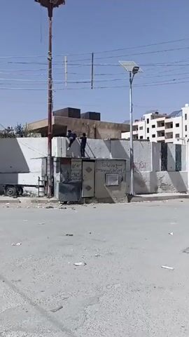 The video shows two young males climbing a gray concrete wall. The wall is topped with large, rough-hewn rectangular blocks. The two males climb onto the wall from the roof of a small, corrugated metal building that is attached to the wall. The building has graffiti or signs written in Arabic on its surface. The first male climbs on top of the wall, and sits down. The second male stands up and briefly gestures toward the seated male. The scene takes place on a sunny day, and a street with some litter on it is visible in the foreground. A taller building can be seen in the background.