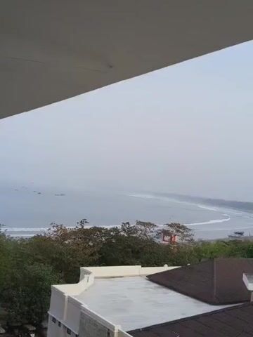 The video captures a wide view of a coastline with a beach and the ocean. In the foreground, there are parts of buildings, including rooftops and walls. The middle ground consists of lush green trees and foliage. The background features a hazy sky, the ocean with gentle waves, and several boats scattered on the water. The overall scene suggests a serene, coastal environment, possibly captured from a higher vantage point. The lighting indicates it might be early morning or late afternoon, given the soft, diffused light. There are no visible people or direct actions occurring within the frame.