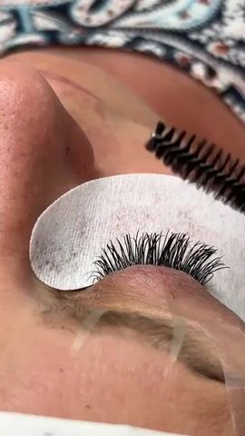 This is a close-up video of an eyelash technician applying eyelash extensions to a client's eye. The client is lying down with her eye closed, and a white cotton pad is placed underneath her lower lashes to protect the skin and catch any stray hairs or product. A small spoolie brush is being used to separate and fan out the client's natural lashes, preparing them for the extensions. The technician's hands and tools are not fully visible, but the focus is on the meticulous process of lash application. The lighting appears to be artificial, suggesting an indoor setting, likely a salon. The client's skin tone is fair, and she is wearing a patterned top or robe, hinting at a professional beauty treatment. The overall atmosphere suggests a calm and precise service being performed. There is no visible text in the media.