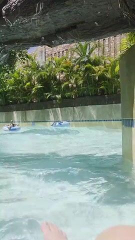 The video shows a view from within a lazy river attraction at a water park. The camera is positioned low in the water, looking forward. In the foreground, the water is choppy, with small waves. Directly above the water is a rock-like overhang, from which water is dripping. Beyond the overhang, the river channel is visible, lined with pale yellow walls. Along the top of the walls is a decorative band of blue and white tiles. On the left side of the river, a person is floating on a blue inner tube. On the right side of the river, a second person is also floating on a blue inner tube, slightly further ahead. Behind the river channel, there is a dense wall of lush green foliage, primarily palm trees and other tropical plants. In the background, a light-colored, multi-story structure with a rough texture is visible, suggesting it is part of the water park's architecture. Sunlight suggests it is daytime.
