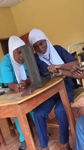 Two women, both wearing white headscarves and medical scrubs, are attending to a patient. One woman, seated and wearing a navy blue scrub set, is taking the patient's blood pressure using a manual sphygmomanometer and a stethoscope. She is intently focused on the equipment and the patient's arm. The other woman, wearing turquoise scrubs, stands beside her, observing the procedure closely. They are at a wooden table on which rests the sphygmomanometer, a cell phone, and other indistinct items. A chair is positioned beside the table and the patient sits in it. The background shows a building with yellow walls and a tile floor.  A metal cot can be seen in the background. The scene appears to be taking place in a medical setting or a clinic.