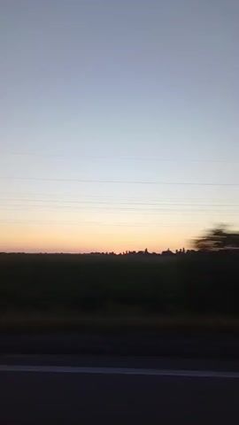 The video shows a view from a moving vehicle. The sky is a gradient of light blue to pale orange, indicating sunrise or sunset. Power lines are visible across the horizon. Below the sky, there is a strip of dark green, likely a field, and then a road or highway. A power pole with multiple crossbars is visible on the left side of the frame. A large, dark-colored truck is seen driving past on the right, its wheels and part of its trailer clearly visible. The truck is moving rapidly, creating motion blur.