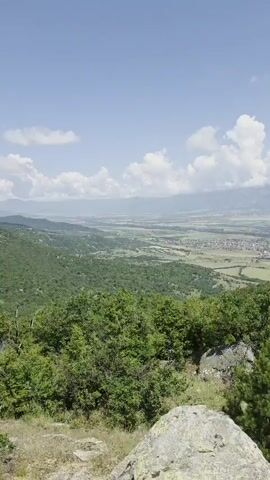 A vertical video of a wide valley landscape. In the foreground, there are large rocks and green bushes. In the middle ground, there are rolling green hills covered in trees. In the background, there is a flat valley with fields and a town. Mountains are visible in the distance under a blue sky with white clouds.