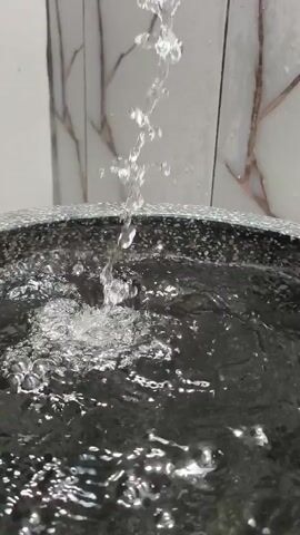 A close-up shot shows water from a fountain falling into a basin. The water is clear and appears to be flowing continuously. The basin is dark-colored and has a textured rim. In the background, a wall with a marble-like pattern is visible. The lighting is bright, and the scene appears to be indoors.