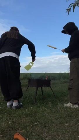Two individuals are outdoors in a grassy area with a blue sky overhead. One person, on the left, is wearing black clothing and a white undershirt. They are leaning over and holding a yellow object, possibly a lid or a tray, as they interact with a charcoal grill. The grill is red and has four legs, and there is smoke and flames emanating from it. The second person, on the right, is wearing a black hooded sweatshirt and olive green cargo pants. They are standing and holding a long stick with something on the end over the grill, appearing to be cooking it. The environment is a field with some trees visible in the background and a knife handle is visible in the foreground.