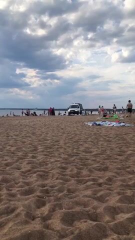 A static shot shows a wide, sandy beach with a light blue lake or ocean in the background, under a cloudy sky. A white pickup truck with a covered bed is parked near the water, facing the camera. Several people are scattered across the beach, with more near the water's edge. Some are standing, others appear to be sitting or lying on towels. A group of people are gathered around an object near the water on the left side of the frame, some in red clothing. The truck's headlights are on.