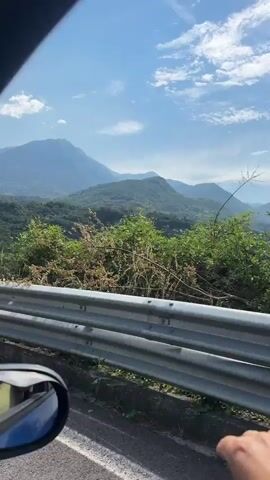 A first-person perspective video shows a hand reaching out over a guardrail. The background is a vast mountain landscape under a blue sky with scattered clouds. The hand moves from right to left, indicating the camera is likely in a moving vehicle. The landscape includes green hills, trees, and distant mountains, with some structures visible on the hillsides. The video is short, capturing a brief moment of the scenic view.
