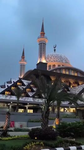 The video presents an exterior view of a large, majestic mosque during dusk or early evening. The sky is a muted blue-grey, and the mosque is illuminated with warm artificial lights, highlighting its architectural details.

The mosque features a prominent central dome, adorned with intricate patterns and topped with a crescent moon finial. Several tall minarets with pointed spires stand around the dome, also brightly lit. The main structure of the mosque displays multiple levels with distinctive rooflines and patterned windows. The lower sections exhibit a unique terraced design with multiple smaller, pointed roofs, suggesting a complex and ornate structure.

In the foreground, lush green vegetation, including several palm trees and shrubs, lines what appears to be a road or pathway. Some of this foliage is also lit. A curb with black and white stripes runs along the bottom edge of the frame. Part of a white vehicle is visible in the lower left corner, indicating the camera is mounted in a moving car.

As the camera pans, a blue rectangular sign comes into view, featuring a white bus icon and the word "STOP," identifying it as a bus stop. Text is also visible on a section of the mosque's facade, reading "PINTU AS-SALAM." The scene is devoid of people, and the atmosphere appears calm under an overcast sky. The overall impression is one of grandeur and tranquility, showcasing a significant religious landmark in Cibinong, Indonesia.