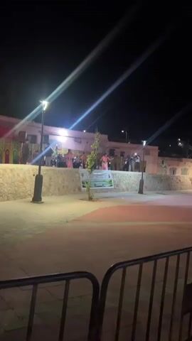 The video shows a woman dancing on a stage at night. The stage is constructed from stone and decorated with plants, and the background is adorned with what appears to be decorative banners. The dancer is wearing a pink dress and a decorative belt. The stage is well-lit by spotlights, and a large speaker can be seen behind the dancer. The surrounding area appears to be a public square or open space, with lampposts and metal barriers visible. Other people can be seen in the background.