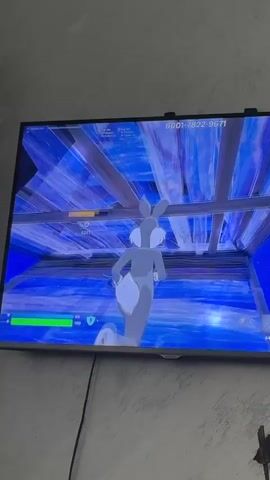 A television screen shows a video game. The character is Bugs Bunny from the Looney Tunes cartoon. He is standing on a structure made of blue panels in a game that appears to be Fortnite. The game information at the top of the screen indicates the game code 6001-7822-9671. Bugs Bunny makes a shooting motion with his hand. The camera is slightly angled upward toward the TV screen. The wall behind the television is painted in a gradient of white to gray. A black cable runs downward from the TV.