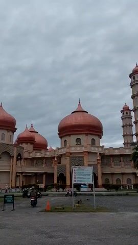 The video shows a large mosque under a cloudy sky. The mosque has a light-colored exterior and multiple domes covered in a copper-colored material. There are two tall minarets with a stacked design. A sign is posted in front of the mosque with various icons. A few people walk around the mosque, and two motorcycles drive on the road in front of the mosque. The parking area in front of the mosque is gray.