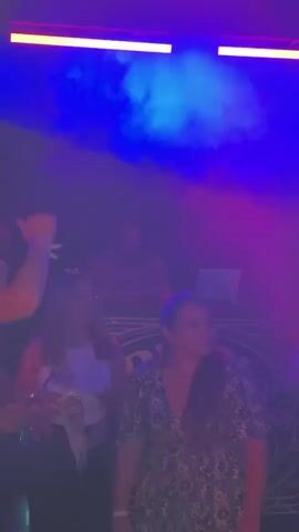 The video shows a nightclub with several people dancing and socializing.  A DJ is visible behind a booth with a laptop. Red lighting and fog effects enhance the nightclub atmosphere. Some people are holding drinks. The camera moves, and it is difficult to keep focus on anyone specific in the crowd.