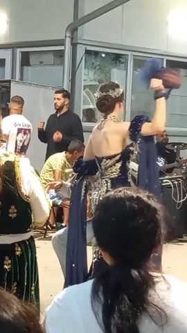A man in a grey suit and green sash dances in the foreground while a woman in an elaborate dark blue dress with gold embellishments dances in the background. They are at an outdoor event with other people watching. There is a band with drums and other instruments behind the dancers. The environment appears to be a covered outdoor space with windows in the background.