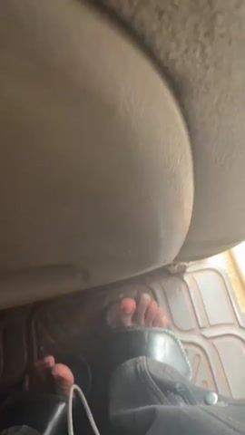 The video shows a first-person perspective of a person's feet inside a vehicle. The feet are bare and appear to be on the floor mat of the car. The person is wearing dark-colored shoes on one foot, while the other foot is bare. The video is shaky, suggesting the vehicle is in motion. The lighting is dim, indicating it may be daytime or evening. The dashboard and steering wheel are visible in the background.