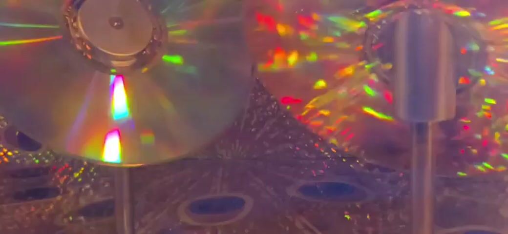 The video shows a close-up, low-angle view of a rotating, metallic object that resembles a kaleidoscope or a spinning toy. The object has a shimmering, iridescent surface with a pattern of circular shapes that look like peacock eyes. There are two vertical silver poles visible in the foreground, one on the left and one on the right, which appear to be part of the structure holding the rotating object. The background is a complex, geometric pattern with a multitude of small, iridescent dots and oval shapes, also resembling peacock feathers. The lighting is dim and artificial, with colorful reflections and highlights dancing across the surfaces of the rotating object and the background. The overall impression is one of an abstract, visually dynamic display, possibly part of an art installation or a decorative element.