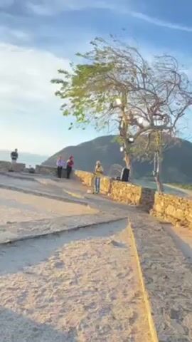 The video shows a sunny outdoor scene with a man playing the saxophone in front of a small crowd. The man, wearing a yellow shirt and blue jeans, stands near a stone wall, with a microphone stand in front of him. Other people stand nearby and watch or look at the scenic view. Behind them is a landscape with a mountain and a view of the ocean under a partly cloudy blue sky. A tree is visible to the right, and the ground is sandy. The camera briefly pans to show more of the surrounding scene.