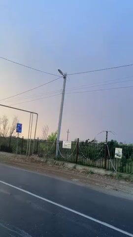 A car drives down a road next to a fence. A rainbow appears in the sky behind the car. The sky is a pale blue and purple. There are power lines overhead. There is a street light on a pole. The fence has barbed wire. There are signs on the fence. Trees and bushes are visible behind the fence. The road appears wet.