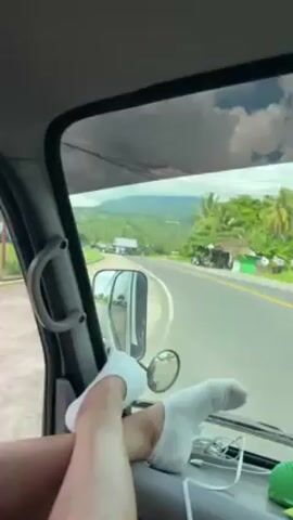 Inside a vehicle moving along a road, a woman with blonde hair, wearing a pink and white striped shirt, a burgundy baseball cap, and denim shorts, has her legs resting on the dashboard. Her feet are clad in white socks. The vehicle passes by fields of lush green vegetation.
