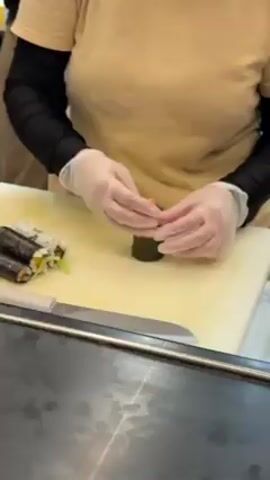 A person wearing a tan t-shirt and black sleeves is making sushi. They are wearing clear plastic gloves. On a white cutting board, they are topping a small, round sushi roll with diced salmon. There is a knife on the cutting board, and another sushi roll to the left. The scene is brightly lit and appears to be in a commercial kitchen.