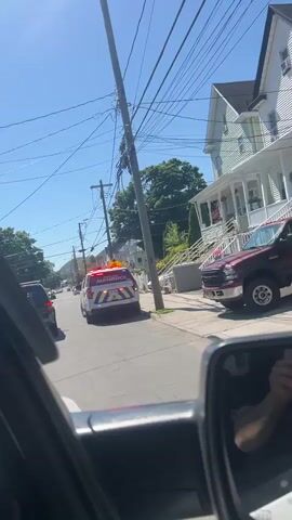 The video, shot from within a vehicle, shows a Rockland Paramedic SUV with flashing lights and a Haverstraw Police SUV following it. The vehicles are moving down a residential street with houses on either side. Utility poles and wires line the street above. The sky is blue and the weather appears sunny. The camera captures the reflection of the person holding the device in the vehicle’s side mirror.