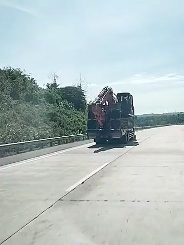 The video shows a truck moving along a highway carrying an excavator. The excavator is dark red and appears to be an Hitachi EX200 model. The truck is dark colored and has several sets of wheels. The highway is smooth and well-maintained with a metal guardrail on the left side.  The landscape is hilly and covered with trees.  The sky is bright, with thin clouds.  The camera is moving along with the truck.
