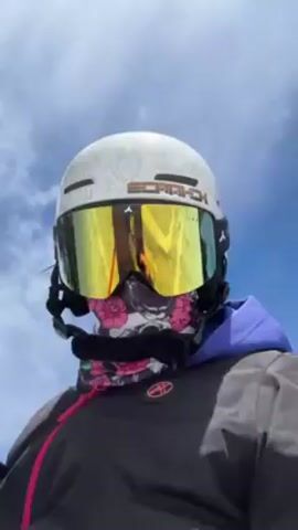 A person wearing a white helmet with the word "SCRATCH" on it, reflective yellow goggles, and a face mask with a skull and floral design is skiing down a snowy slope. The person is wearing a dark jacket with a pink zipper. Trees are visible in the background. The sky is blue with some clouds. The person appears to be filming with a camera pointed at themselves. They are holding ski poles.