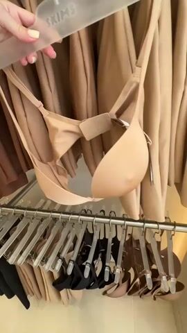 A person's hand with light yellow nail polish removes a beige bra on a clear SKIMS hanger from a clothing rack. The rack contains other bras in varying shades of beige, brown, and black. Behind the rack are garments, presumably of the same material and color palette as the bras. The environment appears to be a retail store.