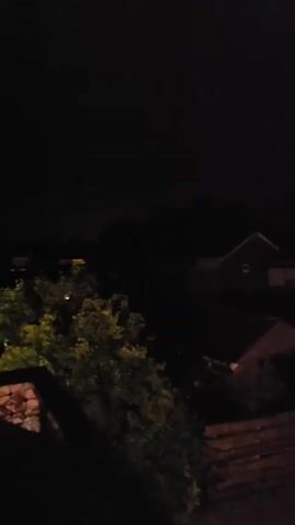 The video shows a dark, possibly nighttime, scene. The camera appears to be elevated, looking down on a residential area with several houses and trees. The sky is visible and periodically flashes with a bright, bluish-purple light, indicating lightning. The scene is largely obscured by darkness, with only glimpses of the structures and foliage illuminated by the flashes and ambient light. The majority of the video is dominated by darkness, with the lightning flashes providing the only significant visual event.