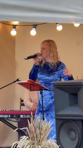 A woman with blonde hair, wearing a blue velvet dress, sings into a microphone on a stage. She stands behind a music stand with an open book. To her left, a red keyboard labeled "Stage 4" is visible. A large black speaker is positioned to her right. Overhead, a string of lights illuminates the scene. The background is a plain, light-colored wall or fabric. The woman's right hand is gesturing as she sings. Another person is partially visible behind her, seated and appearing to play the keyboard. Dried grasses are in the foreground.