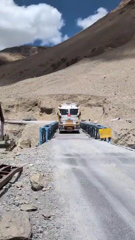 A truck, identified by the "TATA" logo on its front, is captured mid-journey on a narrow bridge. The truck is white and yellow, with various decorations on its roof. The bridge itself is constructed with blue metal railings and a grey asphalt surface. Surrounding the bridge, the landscape is characterized by arid, mountainous terrain with rocky slopes and sparse vegetation. The sky above is partly cloudy, with patches of blue sky visible between white clouds. The video focuses on the truck's slow progress across the bridge, highlighting the challenging environment of the mountainous region.