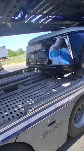 The video shows a car carrier truck on the road. The truck is a multi-level vehicle transporter, and it appears to be carrying new cars. A blue vehicle, possibly a Cadillac Lyriq, is secured on the upper deck of the trailer. The lower deck is metal with a grid pattern. The side of the trailer has the word "Lohr" on it. Other semi-trucks can be seen in the background. The weather appears to be sunny and bright.