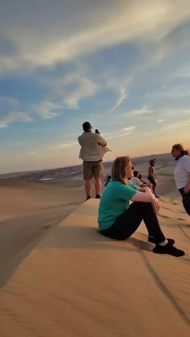 The video depicts a group of people gathered on sand dunes during a sunset. Several individuals are standing or sitting on the dunes, some taking photos with their phones. A red off-road vehicle is parked on the sand, and people are interacting with it. The environment consists of rolling sand dunes under a sky with colorful sunset hues and scattered clouds. The overall setting suggests a recreational or tourist activity.