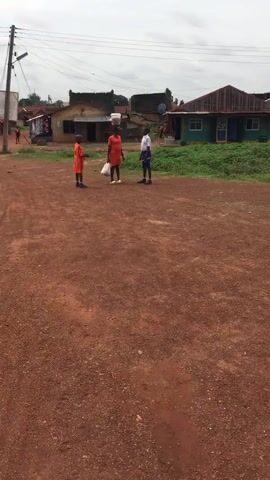 Three children are standing on a dirt road in a rural area. The child in the middle is wearing an orange dress and carrying a plastic bag in one hand, with a white bucket balanced on their head. The other two children are wearing school uniforms, one orange and one white and blue. The background includes small buildings, a power pole, and some greenery. The sky is overcast.