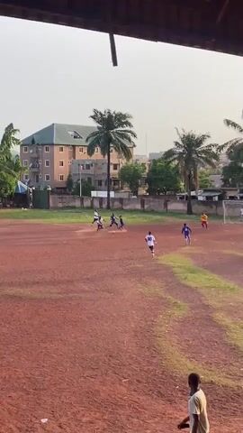 The video depicts a soccer game on a red dirt field with palm trees and buildings in the background. Players in blue and white uniforms are seen in action, with a goalkeeper in a yellow jersey guarding the net. A player in white scores a goal, prompting cheering from onlookers. The camera then shifts to spectators under a covered seating area. Two men, one shirtless and another with dreadlocks and a green tank top, are seen walking up the stairs. The video concludes with a man in a blue and red Adidas tracksuit gesturing while holding a phone and cup. The overall atmosphere suggests a lively, local sports event.