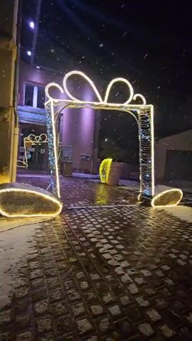 The video shows a nighttime scene in a snow-covered town square decorated for Christmas. A large, illuminated gift box structure stands at the entrance to the square. A cut-out of a reindeer and a Christmas tree stand on the snowy ground. A large Christmas tree, decorated with white and gold lights, is also visible. Snow falls lightly throughout the scene.  The ground is covered with snow and consists of a cobblestone-like pattern. Buildings are seen in the background, and other trees are decorated with lights.  The scene is illuminated by Christmas lights and street lights.