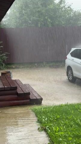 The video shows a heavy downpour of rain in a yard. Water is streaming down and pooling on the ground, creating a wet and slippery surface. A white SUV is parked in the driveway, partially obscured by the rain. To the left, there are wooden steps leading up to a house, also wet from the rain. The yard is surrounded by a dark brown fence, and green trees are visible in the background, their leaves wet and glistening. The overall scene is one of a strong rainstorm impacting an outdoor residential area.