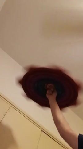 The video shows a person’s arm and clenched fist reaching upwards towards what appears to be a swirling, distorted image of a ceiling. The image has concentric rings of color, predominantly red and darker shades towards the center, giving a blurred and swirling effect. The arm is fair-skinned, and the hand is clenched in a fist. The background is a plain, light-colored wall and a section of what appears to be a wooden door or cabinet. The angle is from a low vantage point, looking up at the ceiling. The focal point is the swirling image and the arm reaching towards it.