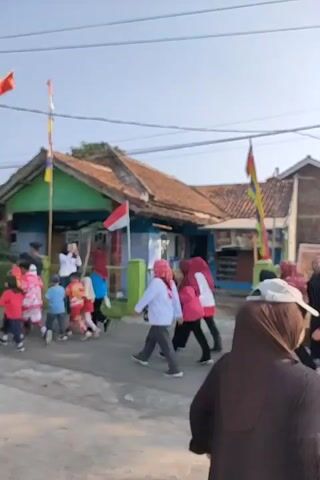 The video shows a group of people, including adults and children, walking down a street. The people are wearing various clothing, many of them are wearing headscarves. There are flags and banners visible in the background, suggesting a celebration or event. The buildings appear to be residential. The sky is clear and the lighting suggests it is daytime.