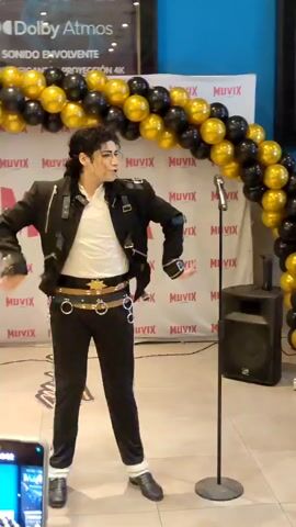 A man dressed as Michael Jackson, wearing a black military-style jacket with silver accents, a white shirt, black pants with silver stripes, and white fur trim on his shoes, stands center frame. He has dark, wavy hair and is gesturing with his hands and head, appearing to be singing or performing. A microphone on a stand is positioned to his right. Behind him, a backdrop features a repeating logo of "MUVIX" in red and white, arched by black and gold balloons. A large screen displaying "Dolby Atmos" and "SONIDO ENVOLVENTE PANTALLA GIGANTE PROYECCIÓN 4K" is visible at the top of the backdrop. A speaker is placed to the right of the microphone stand. The floor is tiled. The scene appears to be an indoor event.