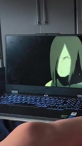 A laptop screen displays an anime scene. The first frame shows a character with long black hair and green eyes against a black background. The second frame displays multiple characters in a stylized environment with red and green energy effects. The laptop is open, with a visible keyboard featuring illuminated blue keys. A person's knee and thigh are visible in the foreground, obscuring the bottom portion of the laptop and the person's lap. The background shows a wall with cabinets.
