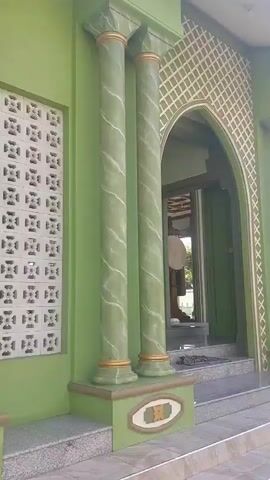 The video shows the exterior of a mosque with green walls and decorative pillars. A staircase leads up to an arched entrance. The pillars are green with marble-like patterns and gold accents. There is a window with a decorative lattice pattern. The overall impression is of a well-maintained religious building.